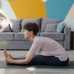Slow motion of attractive curly-haired woman doing yoga at home bending to legs sitting on mat in cozy apartment enjoying healthy activity. People and lifestyle concept.